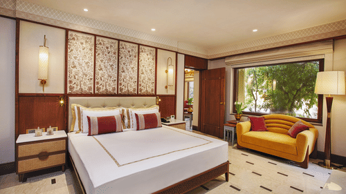 Beautifully designed bedroom with a king size bed, floral accents, a yellow sofa, large window overlooking greenery, and stylish decor - Heritage Village Resorts & Spa, Manesar.