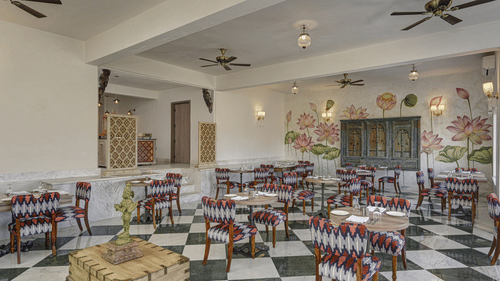 Overview of a brightly lit restaurant interior with a black and white checkered floor, floral-themed wall art, and dark wood furniture at Khas Mahal Suites, Udaipur.