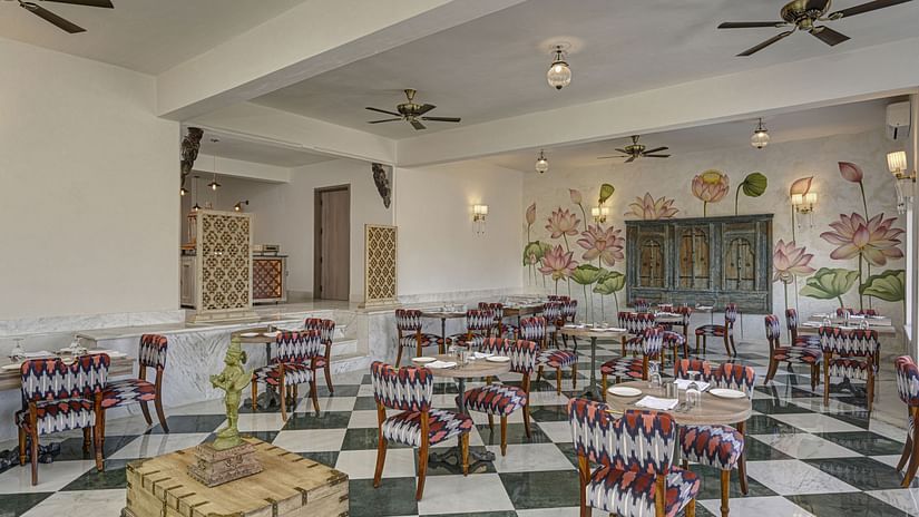 Overview of a brightly lit restaurant interior with a black and white checkered floor, floral-themed wall art, and dark wood furniture at Khas Mahal by Fateh Collection, Udaipur.
