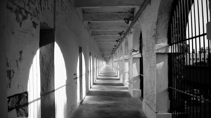 A black and white photograph shows a long, empty prison corridor with arched walkways and barred windows.