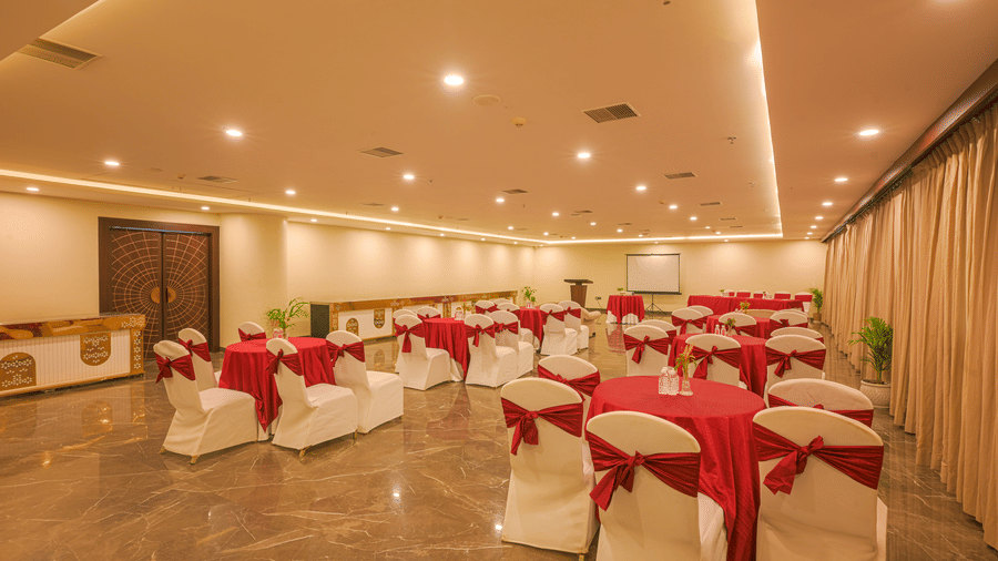 Banquet hall arranged with round tables, white and red covered chairs, floral centrepieces and warm ceiling lights.