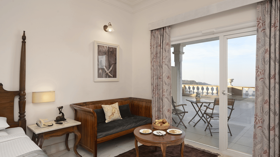 Deluxe Room with Balcony at Fateh Safari Resort, Kumbhalgarh featuring a seating area with sofa, centre table, a corner of the bed, bedside table, and chairs and tables on the balcony.