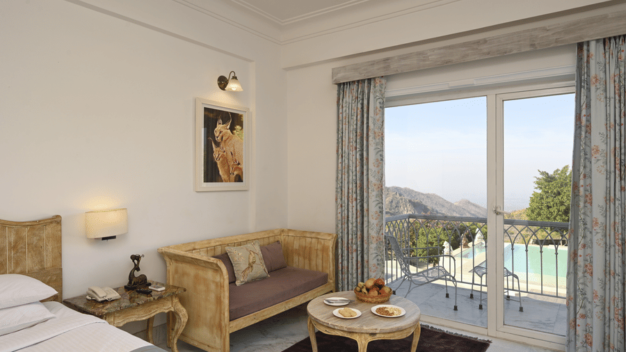 Seating area of Superior Room with Balcony at Fateh Safari Resort, Kumbhalgarh featuring a sofa with centre table next to a bed, and a plunge pool in the balcony with a view of the green hills.