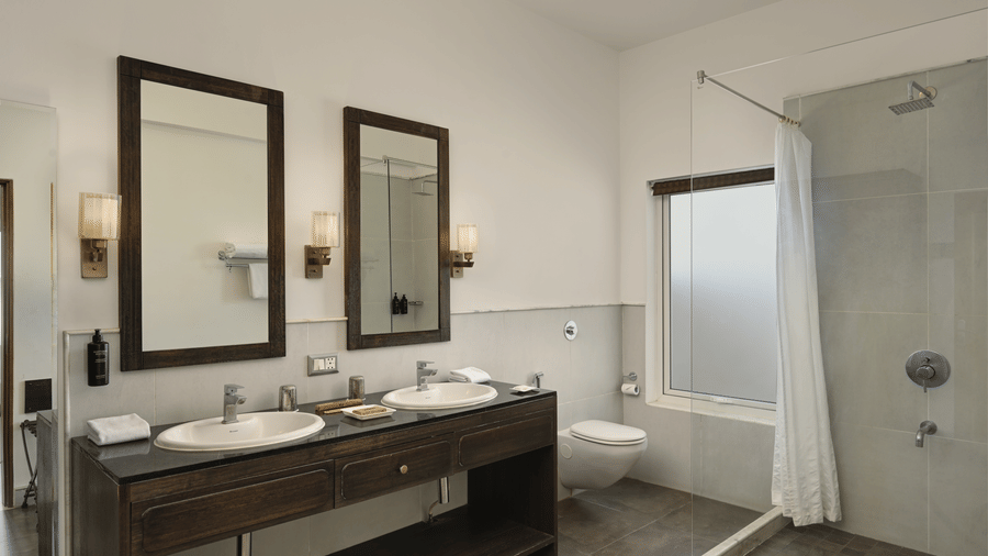 Bathroom of Plunge Pool Suite in Fateh Safari Suites featuring sinks with faucet and mirror attached on the walls, shower screen, and other toiletries in different corner of the room.