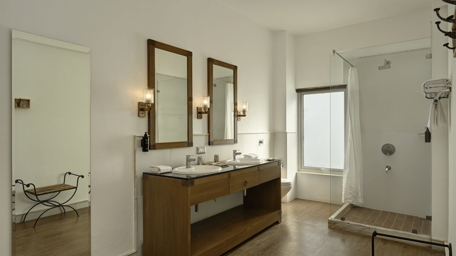 Bathroom of Plunge Pool Suite with Jacuzzi at Fateh Safari Suites, Kumbhalgarh featuring a shower head with glass screen, sinks with faucets and mirror attached on the wall, and other toiletries.
