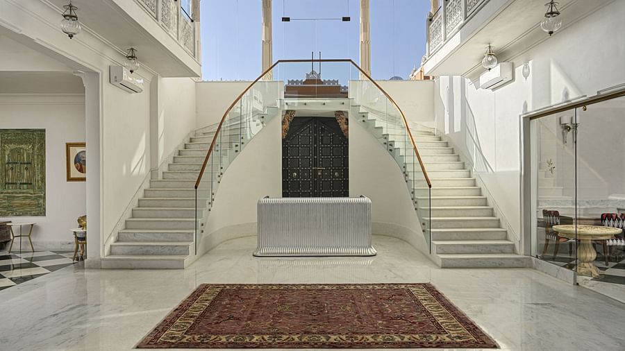 Interior view of a reception at Khas Mahal by Fateh Collection featuring a symmetrical, double staircase with white risers and stringers, glass railings, a dark central door, a brown rug on the floor, and natural light entering from a high window.