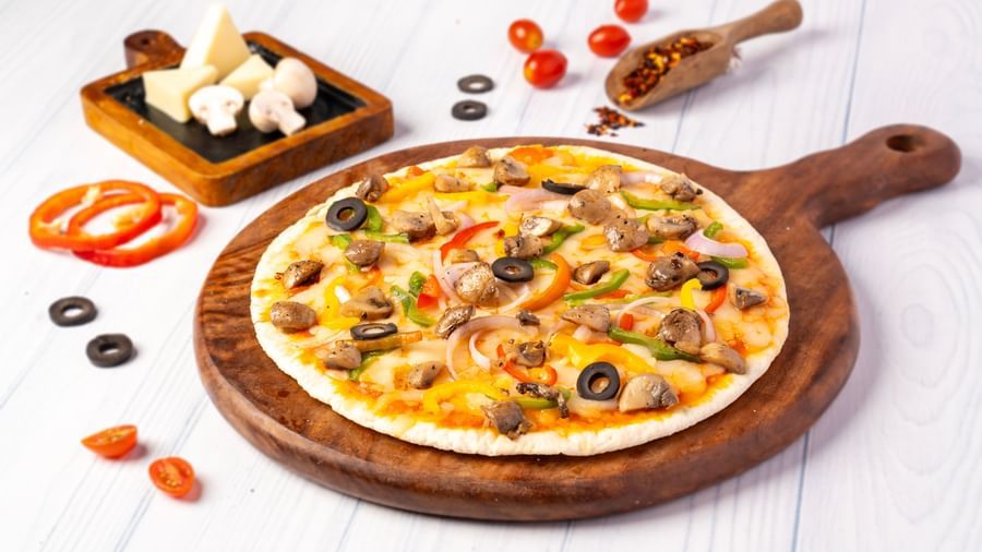A round pizza topped with meat, olives, and vegetables is on a wooden board, surrounded by fresh tomatoes, cheese wedges, and spices.