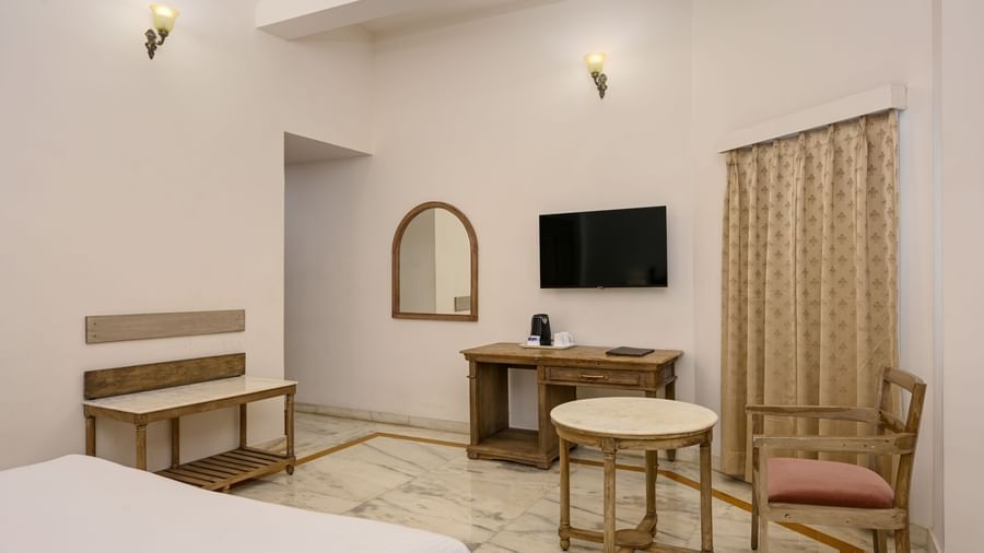 A room at Ram Pratap Palace in Udaipur, featuring a bed, a wall-mounted TV, a wooden desk, a bench, a small mirror, and a coffee table with a chair.