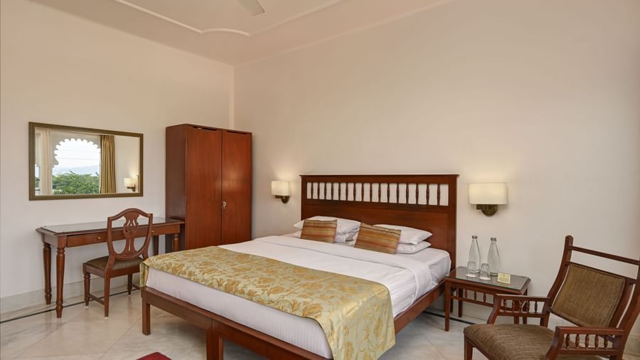 A spacious guest room at Ram Pratap Palace with a large bed, two chairs, a wardrobe, a desk, a mirror, and two bedside tables with lamps.