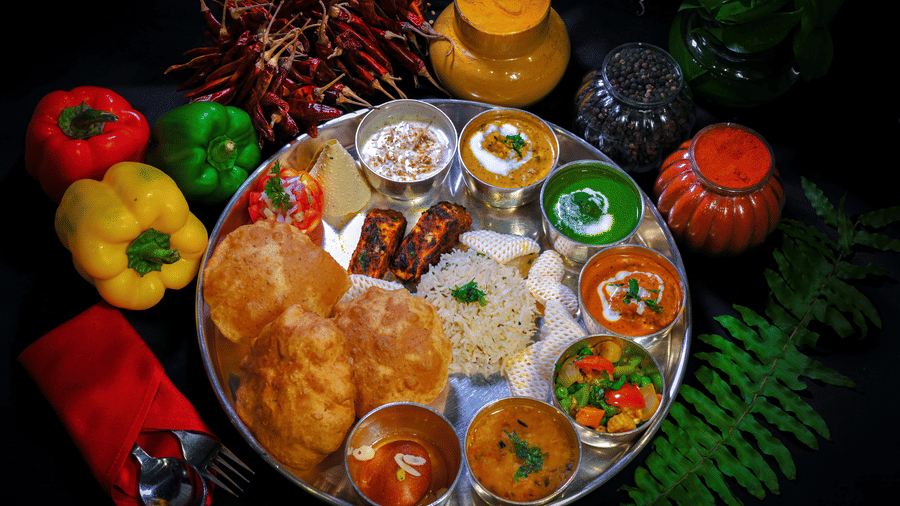 A thali with multiple dishes placed in small cups kept on a plate with capsicums in the backdrop.
