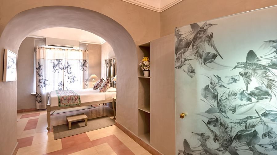 Hallway with an arched opening to a room with a daybed and patterned curtains, a cabinet with shelves, and a decorated glass door at Tijara Fort-Palace, Alwar.