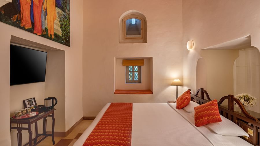 A bedroom with a bed, a table, a television, a window, and wall art in Tijara Fort-Palace - 19th Century, Alwar.