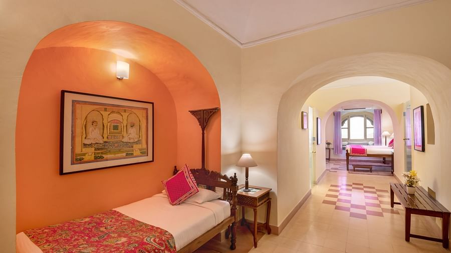 Hotel room with a single bed in an orange alcove with artwork, a bedside table with a lamp, and a view through an archway to a hallway and another bedroom at Tijara Fort-Palace, Alwar.
