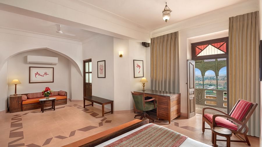 Room with bed, wooden frame, patterned runner, furniture including sofa, desk, chairs, and table, curtains, artwork, decorative doorway, and patterned floor - Tijara Fort-Palace - 19th Century, Alwar.