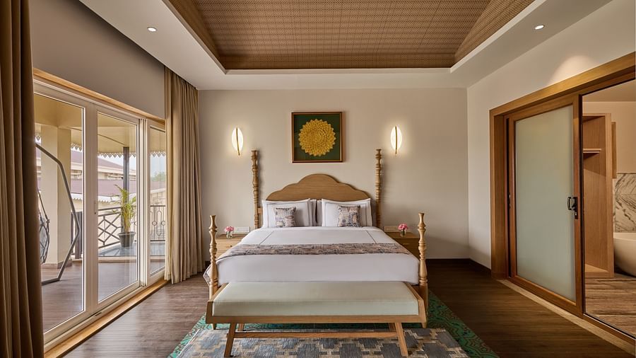 Elegant hotel bedroom with a wooden bed frame, white bedding, decorative rug, sliding glass doors leading to a balcony, and warm ambient lighting - Voyage The Solitaire Bliss.