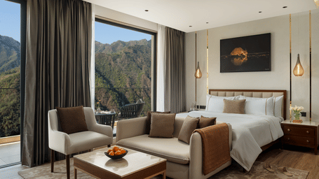 A bright hotel room with a sofa set, double bed, and large windows revealing a beautiful mountain landscape.