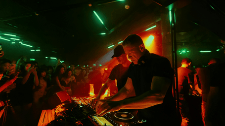 An overview of a club with the DJ mixing songs on a turntable and people dancing next to him.