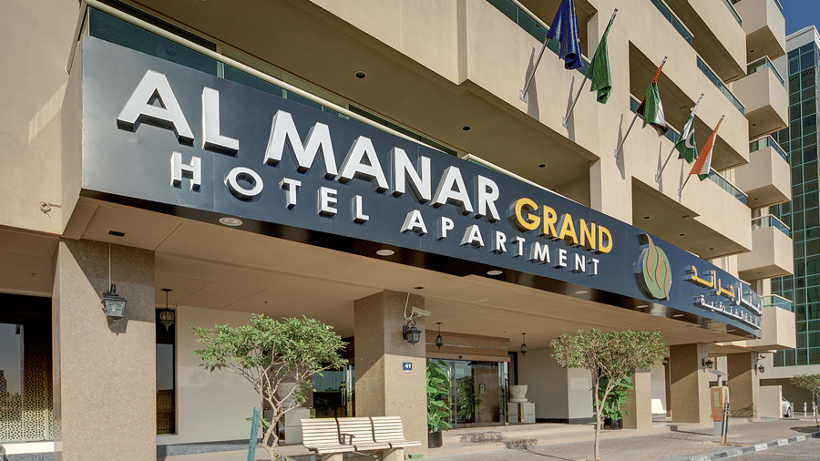 The entrance of the hotel building showing a large sign, flags, and small trees outside the building - Al Manar Grand Hotel Apartment, Bur Dubai