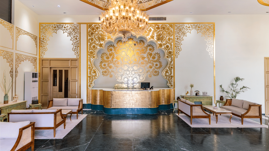 A reception area at Beelwa Palace, Jaipur, with chandelier, decorative wall detailing, seating arrangements along the sides, and polished dark flooring.