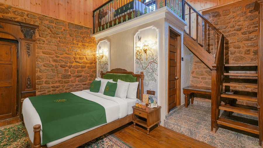 The executive-suite at BluSalzz Terrah Hills Resort, Dalhousie, featuring a bed with a green and white duvet, wooden flooring, exposed brick walls, and a wooden staircase leading to an upper level.