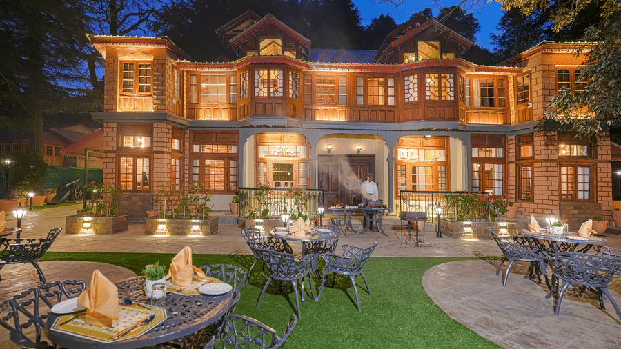 The facade of BluSalzz Terrah Hills Resort, Dalhousie, illuminated with lights, a man standing near the grill, huge trees in the backdrop, and an outdoor seating area on a green lawn.