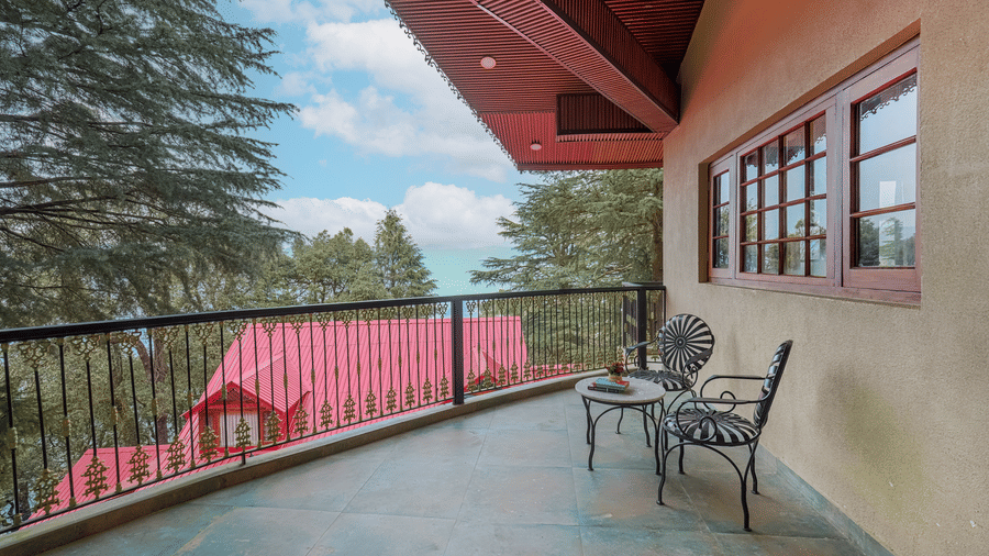 The balcony of BluSalzz Terrah Hills Resort, Dalhousie, with a railing, two chairs, a table, and forested hills in the side.