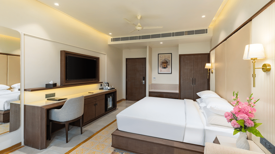 An interior of a room at EsthereaRaj Leela, Ranakpur, with a king size bed, a desk chair, a built-in wardrobe, and a flat-screen television on a wooden unit.