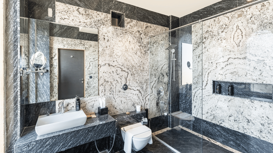 A large bathroom featuring grey-veined marble walls, a walk-in shower with a glass door, and a modern vanity area at EsthereaRaj Leela, Ranakpur.