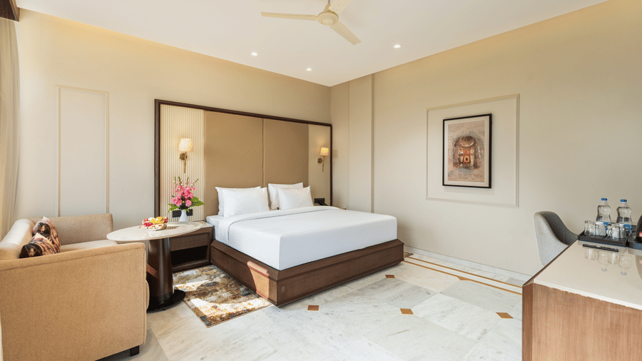 A bedroom at EsthereaRaj Leela, Ranakpur, with a king size bed, neutral-toned headboard, white tiled flooring, a wall art, and wooden furniture.