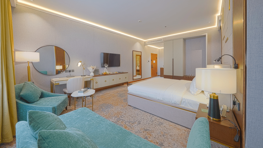 Deluxe King bedroom corner view showcasing the plush bed facing the wall-mounted TV, the work desk situated beside an upholstered couch | Grand Iva Hotel, Abha