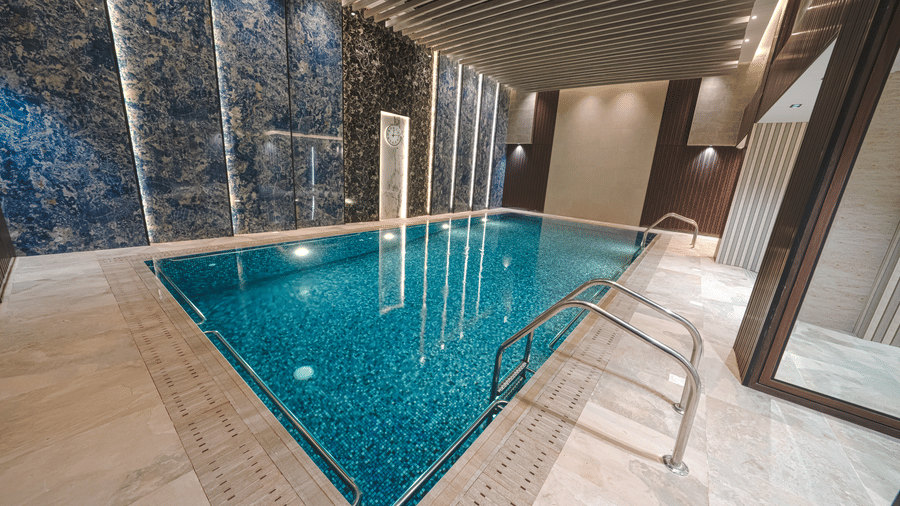 Indoor swimming pool at Grand Iva Hotel, Abha, seen with a clean and spacious deck, a panelled wall behind it with a clock on it.