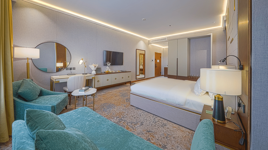 Junior Suite bedroom corner view showcasing the plush bed facing the wall-mounted TV, the work desk situated beside an upholstered couch | Grand Iva Hotel, Abha