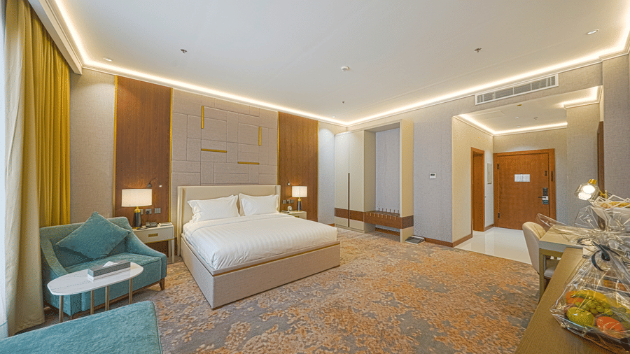 Corner view of the Junior Suite's bedroom at Grand Iva Hotel, Abha, featuring modern wooden panelling, upholstered chair, and ample lighting.