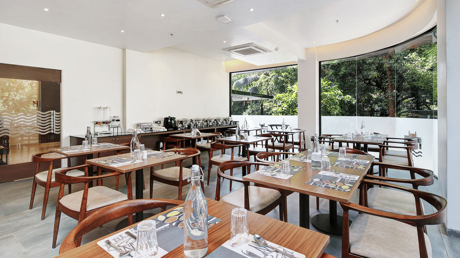 Modern restaurant dining area with wooden tables and chairs, natural light, and buffet counter at Hotel Mumbai House, Ghansoli.