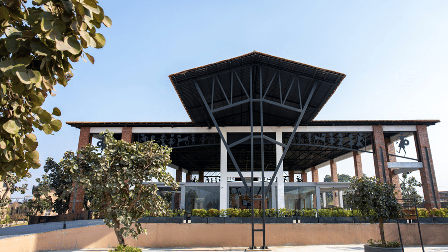 A front-facing exterior view featuring a modern building with a symmetrical dark roof structure, prominent brick pillars, and a low boundary wall with plants at Hotel Sonar Bangla, Ajodhya Hills.