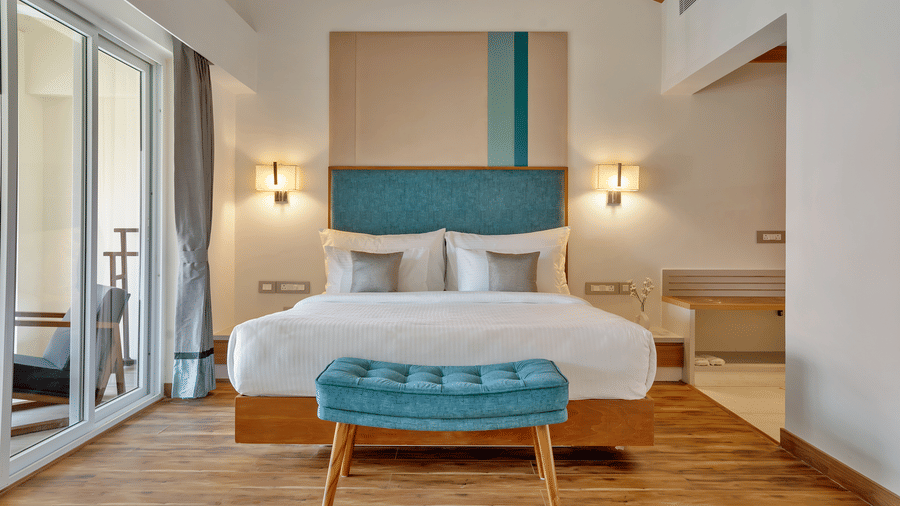 Bedroom of Suite at Mango Hill, Velankanni with a bed, bench at the foot of the bed, wall lamps, and glass door.