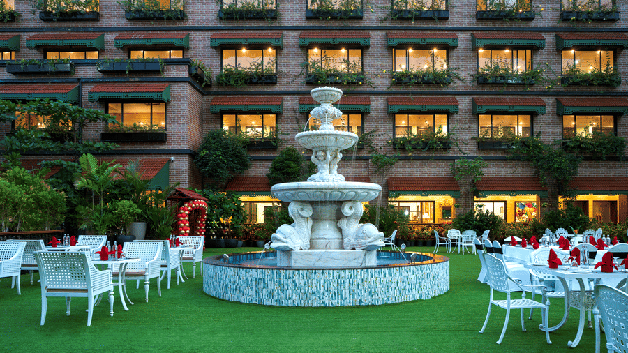 Outdoor dining area on a manicured lawn next to MAYFAIR Bay Resort, Paradeep's facade, set up with white tables and a fountain in the middle.