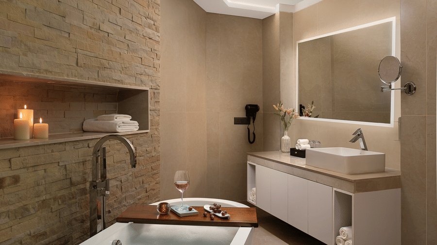 A cosy bathroom setting with a bathtub, a white vanity unit, and glowing scented candles against a textured stone wall.