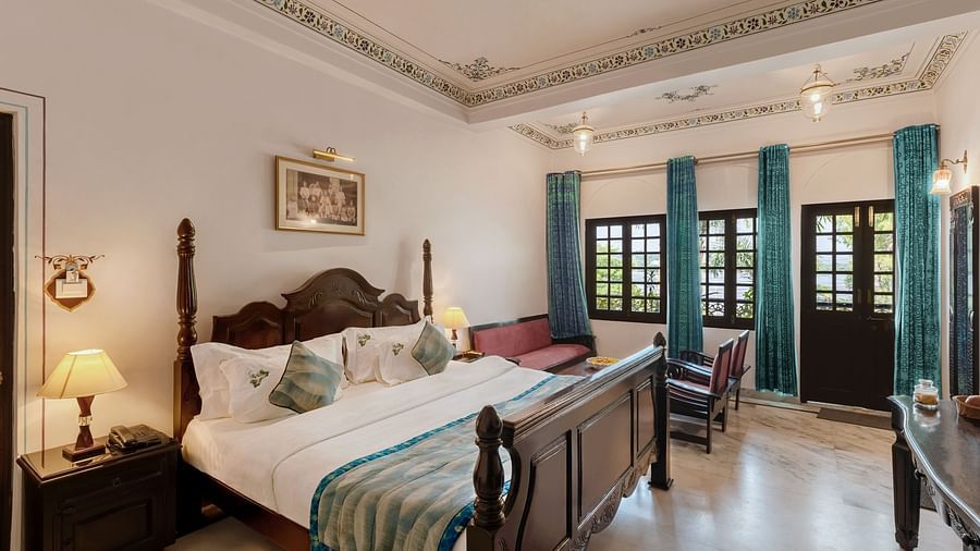 A spacious bedroom at Panna Vilas Palace, featuring a bedside table with a lamp and a series of windows to the side, with curtains pulled back | Panna Vilas Udaipur