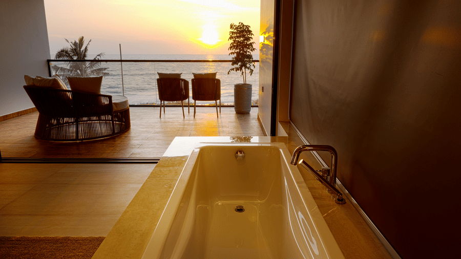 Sunset view bathtub at SUKAVI in a modern bathroom with a balcony view showing the sun setting in the distance.