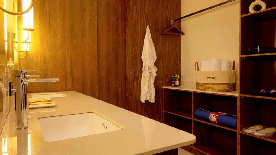 Bathroom at SUKAVI with a floating dark wood vanity, 2 sinks, mirrors with vertical lights, and a white bathrobe hanging on a hook.