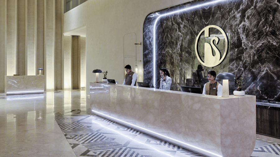 The reception lobby at Swosti Premium Beach Resorts, Puri, featuring a long, illuminated marble counter, a wall with a swan logo, and staff members working behind the desk.