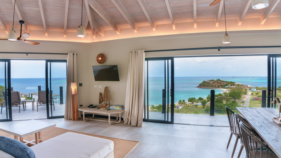 An open-plan living and dining area with a dining table, sofas, ceiling fans, and wide glass doors offering views of the swimming pool, sea, and coastline at Suite 35, Tamarind Hills Resort and Villas.