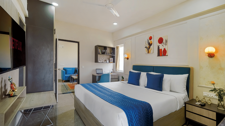 A room at Urban Suites by BluSalzz, Jaipur, with a large bed featuring white and blue bedding, a headboard, a bedside lamp, and a door giving way to a living room.