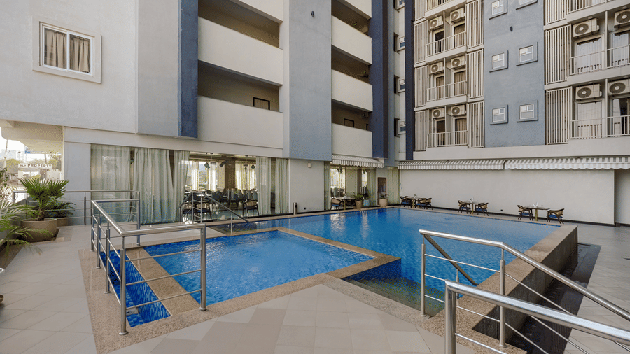 An outdoor swimming pool, showing a shallower section and a deeper section, surrounded by railings and part of a building structure.