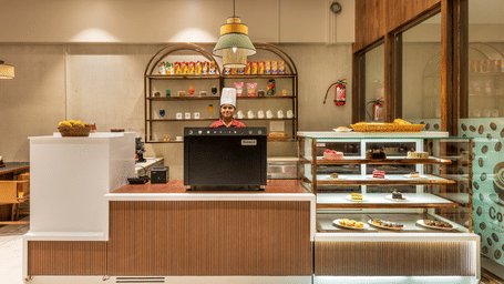 A modern cafe service counter at L Café with a glass display case for pastries and snacks, complemented by warm wood panelling and minimalist decor – Skkylight Lords Inn, Nanded