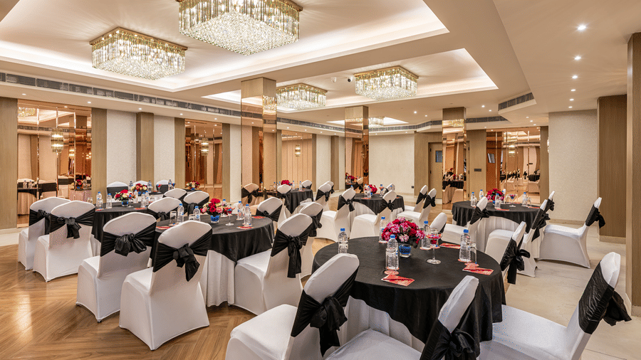 An elegant spacious hall with round tables and white chairs adorned with black fabric, featuring a decorative ceiling with hanging lights and mirrored wall accents at Lords Eco Inn Dehradun.
