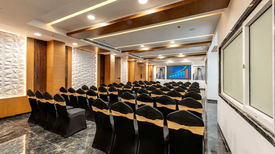 Banquet room with rows of black chairs facing a presentation area, adjacent to large glass windows.