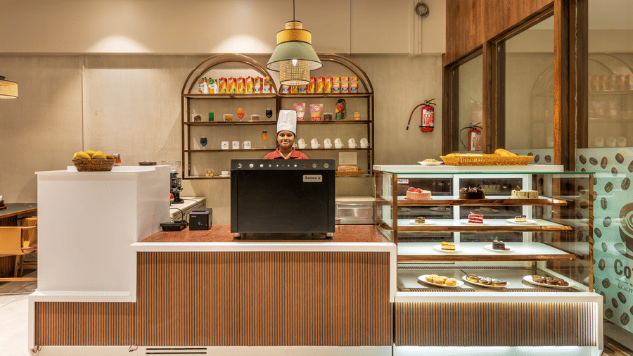 A modern cafe service counter at L Café with a glass display case for pastries and snacks, complemented by warm wood panelling and minimalist decor – Skkylight Lords Inn, Nanded