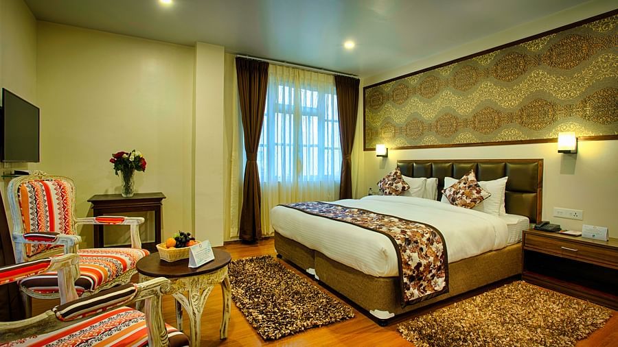 full view of suite room showcasing a king size bed, 2 chairs and a teapoy, a television and much more along with curtained windows - the aryan regency